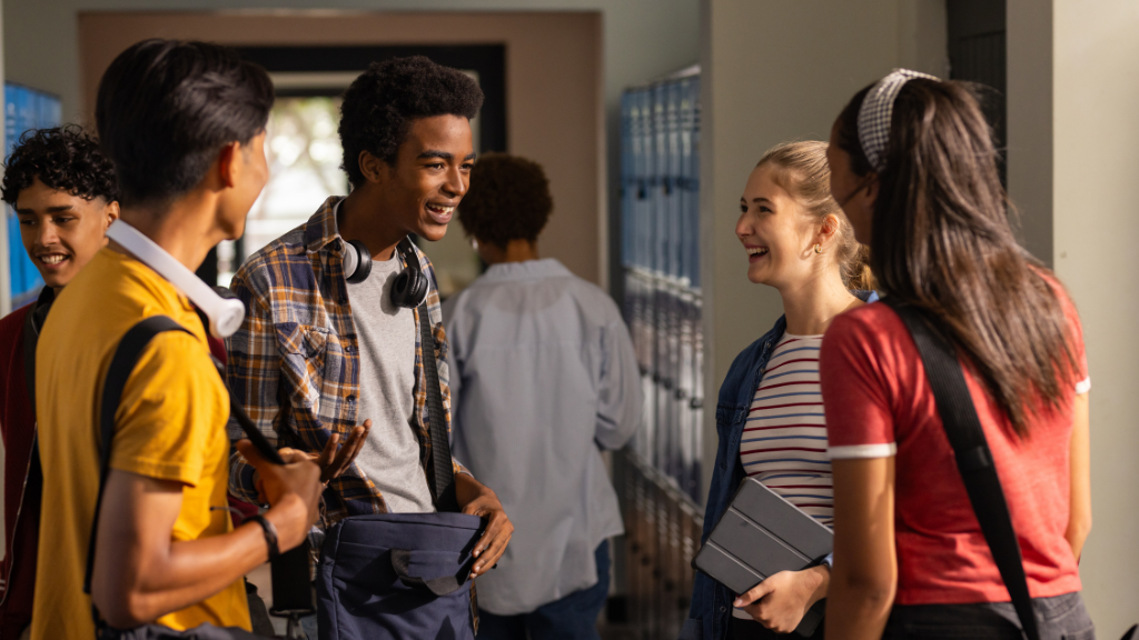 A group of high school students stand near school lockers, talking and smiling together between classes.